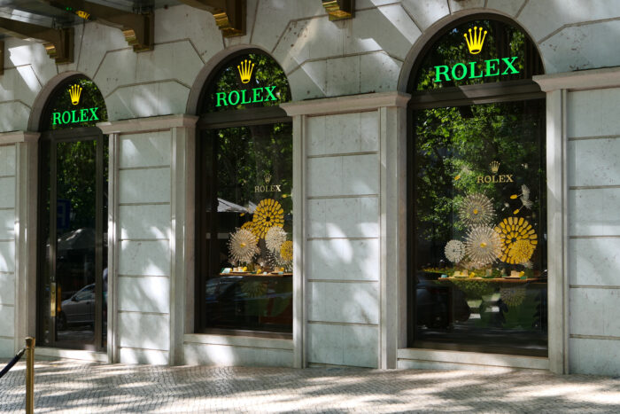A Rolex storefront with elegant arched windows and glowing green-and-gold branding sits along a tree-lined street. Reflections of lush greenery mingle with artistic window displays, creating a sophisticated scene that highlights the brand’s refined retail presence.