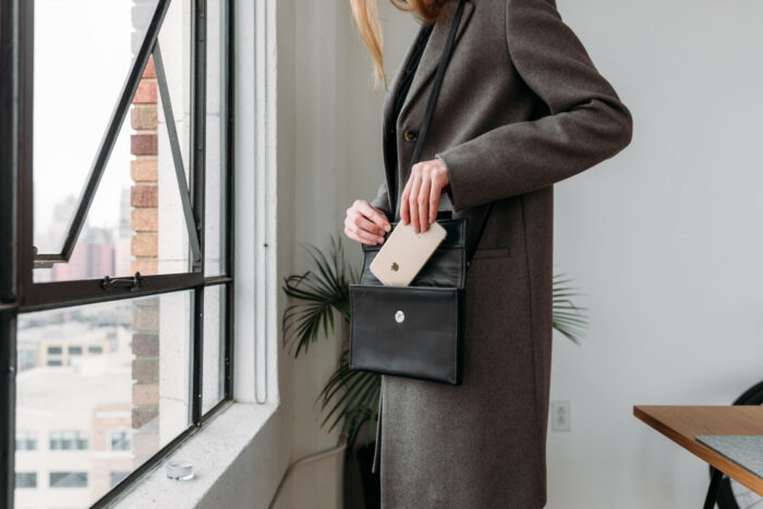 A person wearing a long gray coat places a smartphone into a black crossbody bag while standing beside a large window in a bright, modern office or apartment, highlighting secure storage and everyday carry essentials.