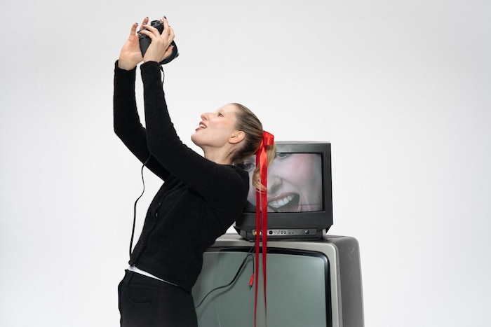 Woman with a camera whose feed is appearing on a CRT behind her