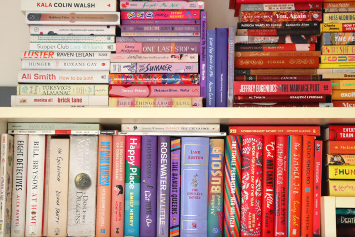 Shelves filled with colorful books arranged both vertically and in stacked piles. The collection includes contemporary fiction, classic literature, and nonfiction, creating a vibrant display of varied book spines in bright reds, blues, purples, yellows, and whites.