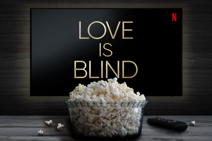 A bowl filled with freshly popped popcorn sits in front of a television displaying the title "Love Is Blind" on Netflix. A TV remote rests on the table beside scattered popcorn kernels, suggesting a relaxed movie or binge-watching setup.
