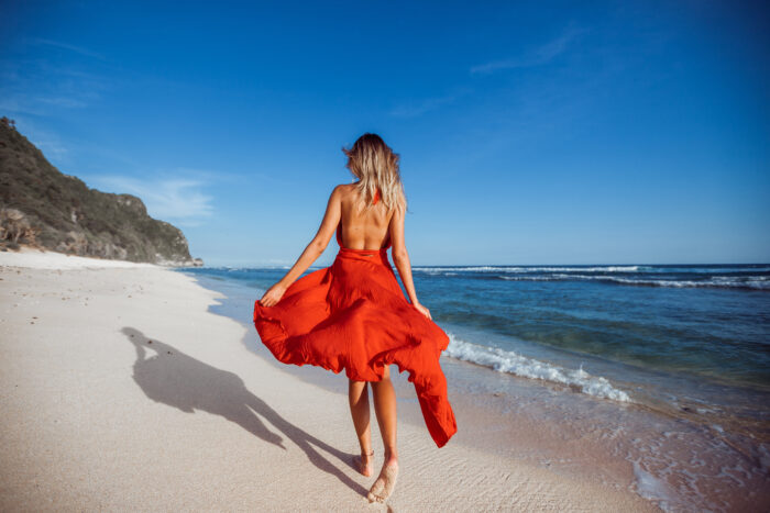 A woman in a flowing backless red dress walks barefoot along a white sand beach, holding the skirt as it billows in the ocean breeze. Viewed from behind, she faces the turquoise water beneath a cloudless blue sky, with rocky cliffs stretching along the shoreline to her left.