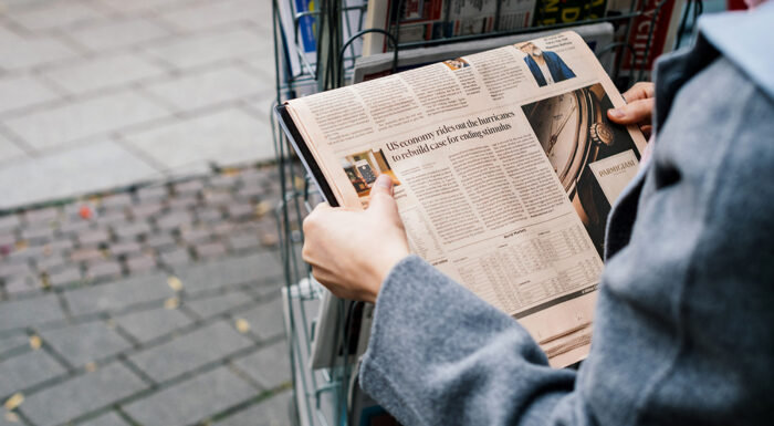 A person wearing a gray coat browses a newspaper at an outdoor newsstand, holding an open copy featuring financial news and a luxury watch advertisement, with rows of newspapers and magazines displayed in the rack behind them.