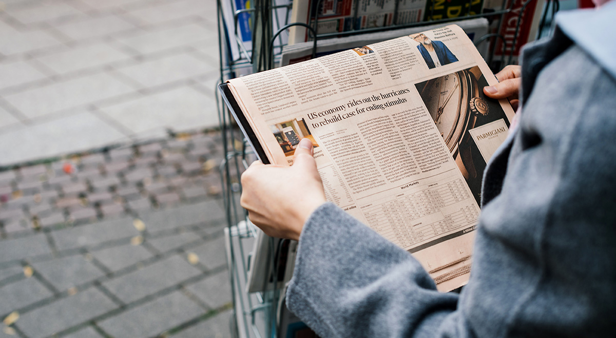 A person wearing a gray coat browses a newspaper at an outdoor newsstand, holding an open copy featuring financial news and a luxury watch advertisement, with rows of newspapers and magazines displayed in the rack behind them.
