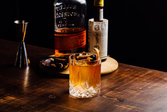 A classic whiskey cocktail served over ice in a cut-crystal rocks glass, garnished with an orange peel, cherry, and cinnamon stick, sits on a wooden bar beside bourbon bottles and bar tools.