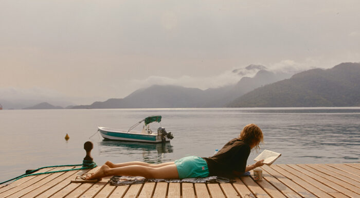 A person lies on a wooden dock reading a book beside a calm lake, with a small motorboat anchored nearby. Mist drifts over tree-covered mountains in the background beneath a soft, overcast sky, creating a peaceful, secluded atmosphere.