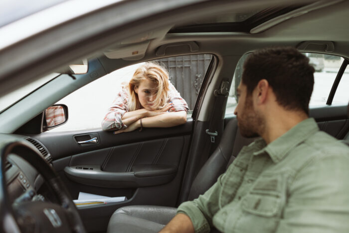 Viewed from inside a parked car, a woman leans on the open passenger-side window with her arms folded, looking down with a somber expression as she speaks to a man seated in the driver's seat. The candid moment suggests a serious or emotional conversation.