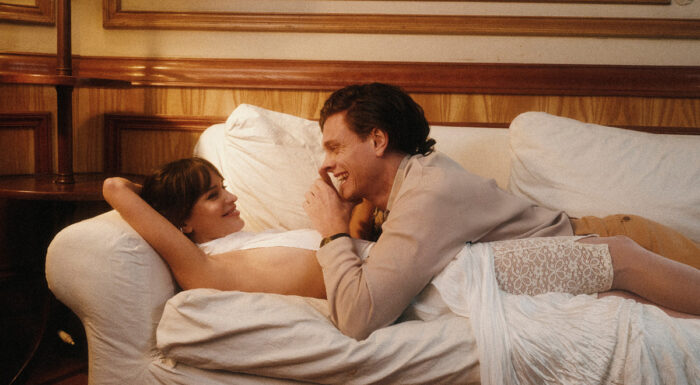A smiling couple reclines on a white sofa in a warmly lit room with wood-paneled walls. The woman, wearing a flowing white dress with lace stockings, gazes up at the man as he leans over her, laughing. Their relaxed posture and affectionate expressions create an intimate, romantic atmosphere.