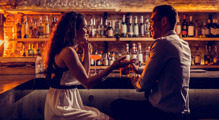 A man and woman sit across from each other at a warmly lit cocktail bar, smiling and engaged in conversation over drinks, with shelves of liquor bottles creating an intimate, inviting atmosphere in the background.