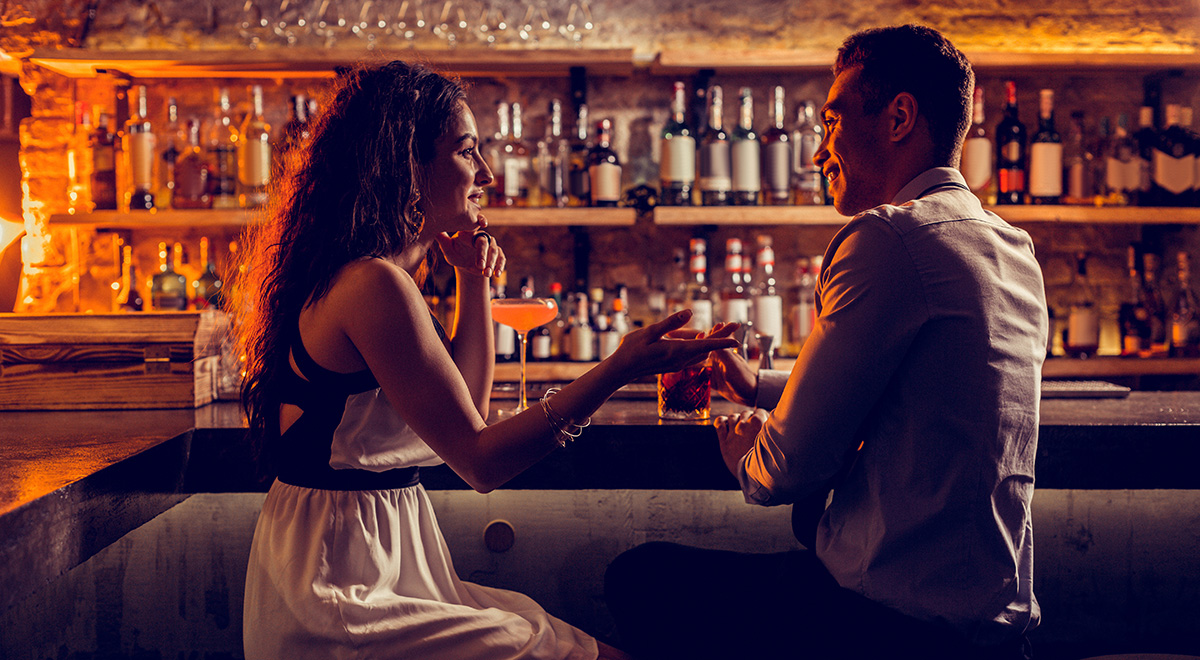A man and woman sit across from each other at a warmly lit cocktail bar, smiling and engaged in conversation over drinks, with shelves of liquor bottles creating an intimate, inviting atmosphere in the background.