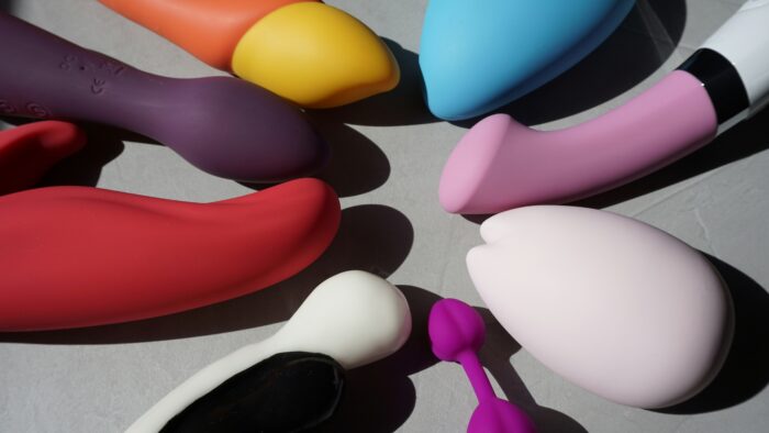 A collection of colorful silicone personal wellness devices and accessories is arranged on a neutral surface. The assortment features a variety of shapes, sizes, and pastel tones, highlighting modern intimate wellness products in a clean, minimalist composition.