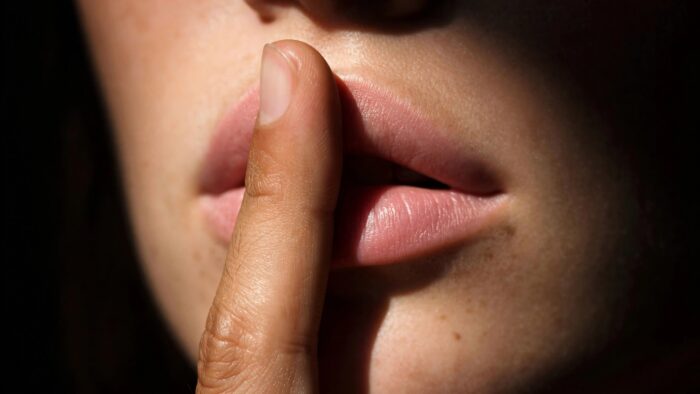 Close-up of a person holding a finger to their lips in a “shh” gesture against a dark background.