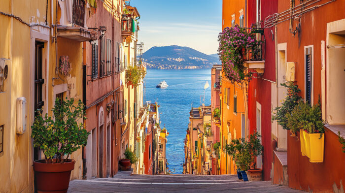 A colorful Italian street lined with warm-toned buildings and flower-filled balconies descends toward the sparkling blue sea, where boats cruise beneath distant coastal hills.