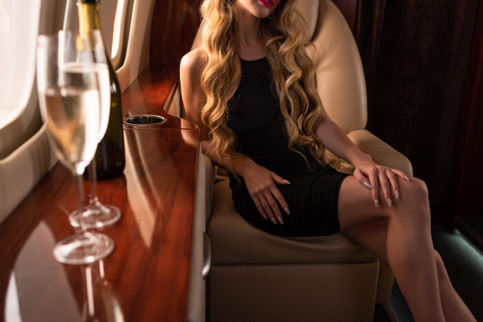 A woman in a black dress relaxes aboard a private jet, with champagne glasses and a bottle displayed beside her in the luxurious cabin.
