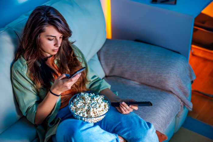 Beautiful young woman relaxing at home at night, having fun sitting on the couch watching TV and typing text message using smart phone
