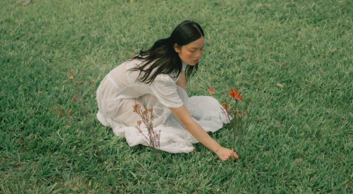 A woman in a flowing white dress sits in a grassy field dotted with orange wildflowers, reaching toward the grass.