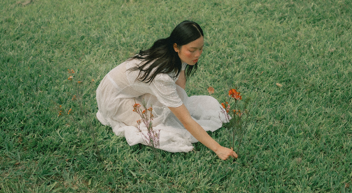 A woman in a flowing white dress sits in a grassy field dotted with orange wildflowers, reaching toward the grass.