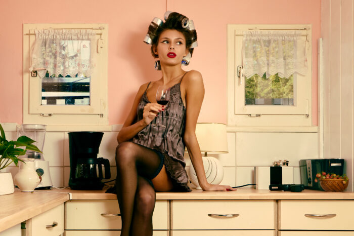 Woman in lingerie and hair rollers sitting on a kitchen counter while holding a glass of red wine in a retro-inspired pink kitchen.