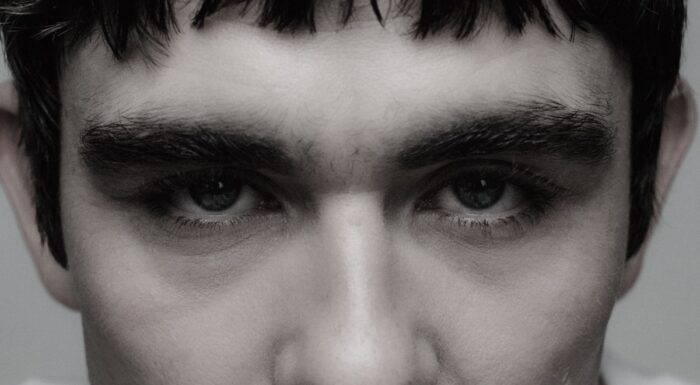 A tightly cropped portrait focuses on a person's blue eyes framed by thick dark eyebrows and short bangs. Soft lighting highlights the skin's natural texture and creates an intense, contemplative gaze.