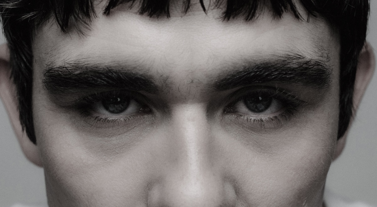 A tightly cropped portrait focuses on a person's blue eyes framed by thick dark eyebrows and short bangs. Soft lighting highlights the skin's natural texture and creates an intense, contemplative gaze.