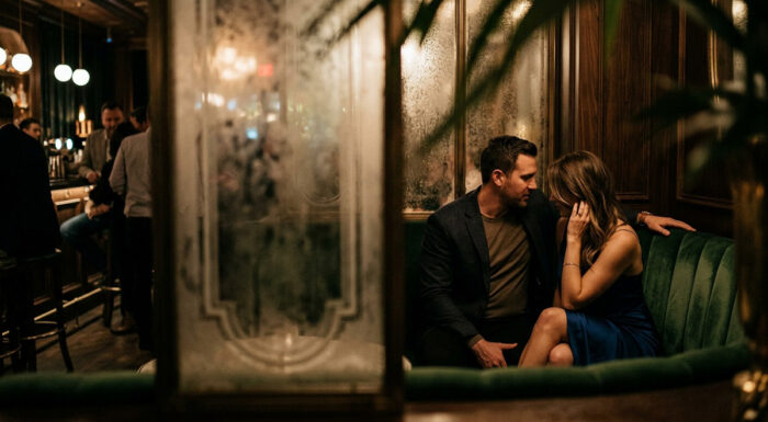 A couple sits closely together in a dimly lit bar, speaking intimately on a green velvet banquette while other patrons gather in the background.