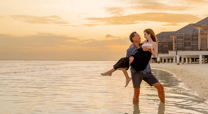 Dana Rosewall and Brandon Wade sharing a playful moment in shallow ocean water at sunset, with overwater villas lining the beach in the background.