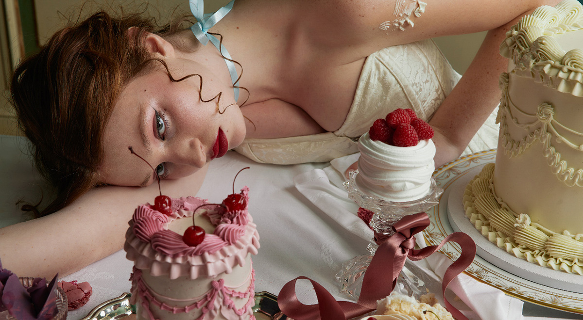 Woman in a white dress reclining across a table surrounded by ornate cakes, cherries, raspberries and ribbons.