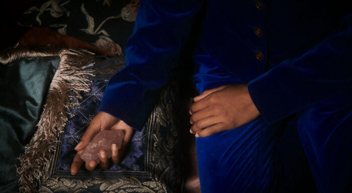 Close-up of a person in a royal blue velvet suit seated on an ornate sofa, holding a rose quartz crystal in one hand.