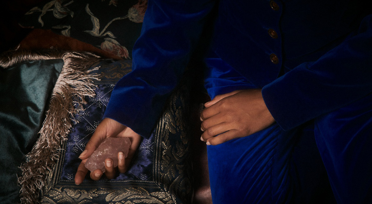 Close-up of a person in a royal blue velvet suit seated on an ornate sofa, holding a rose quartz crystal in one hand.