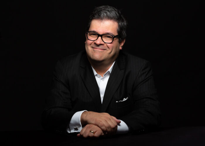 Michael Spremulli, wearing glasses and a dark pinstripe suit, poses against a black background.