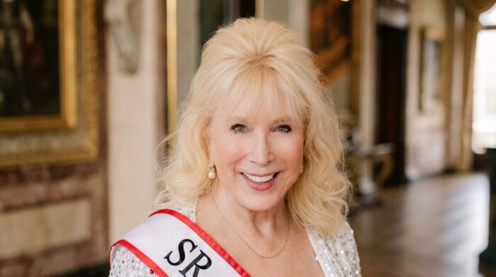 Human behavior expert Dr. Lillian Glass smiles while wearing a white sash with red trim in an elegant interior. AI disclosure: AI-assisted image; original portrait reformatted from vertical to landscape.